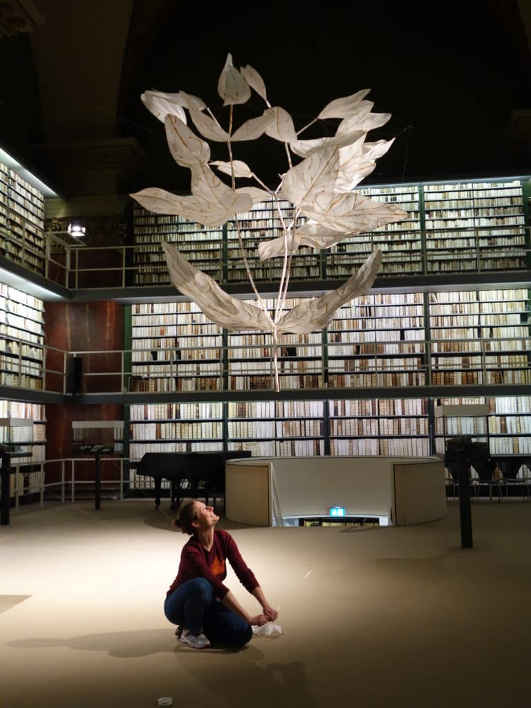 Eine Frau kniet auf dem Boden. Über ihr schwebt eine Pflanze aus Papier. Im Hintergrund sind Bücherbestände in der Augusteerhalle zu sehen.
