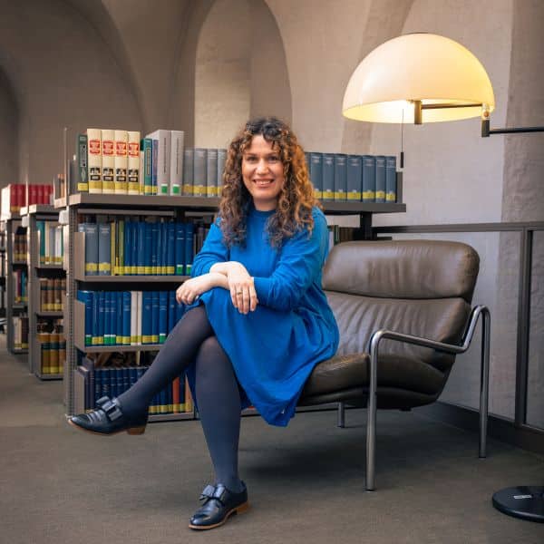 Dr. Irena Fliter auf der Empore des Zeughauses in der Herzog August Bibliothek Wolfenbüttel. Sie sitzt in einem tiefen Ledersessel. Hinter ihr sind mehrere Bücherregale zu sehen. Auf der rechten Seite sieht man eine Leselampe.