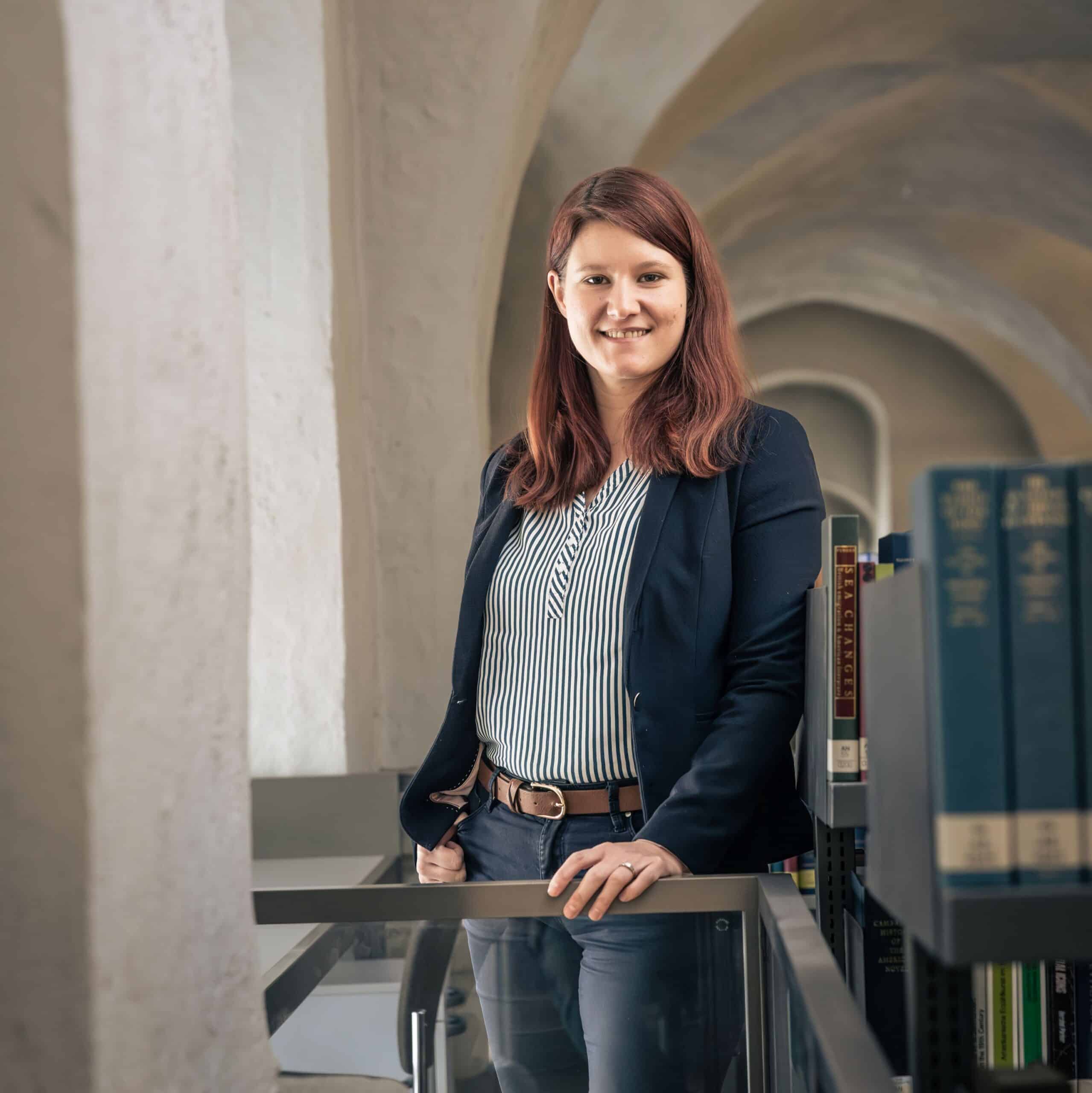 Dr. Anna Lingnau auf der Empore im Zeughaus. Sie lehnt an einem Geländer und schaut in die Kamera. Hinter ihr erstreckt sich das Deckengewölbe.