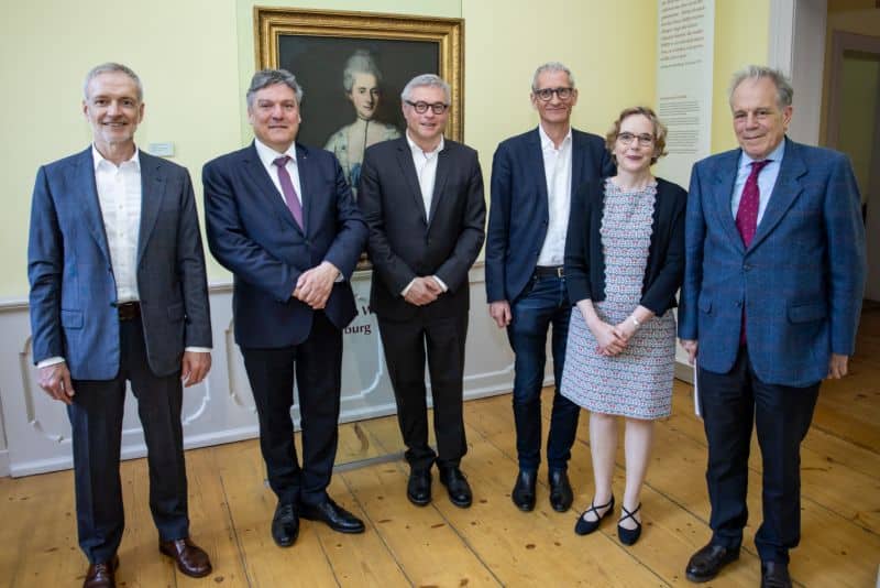 Gruppenbild mit sechs Personen in einem Raum. Im Hintergrund hängt ein gerahmtes Ölgemälde, das Eva König zeigt.